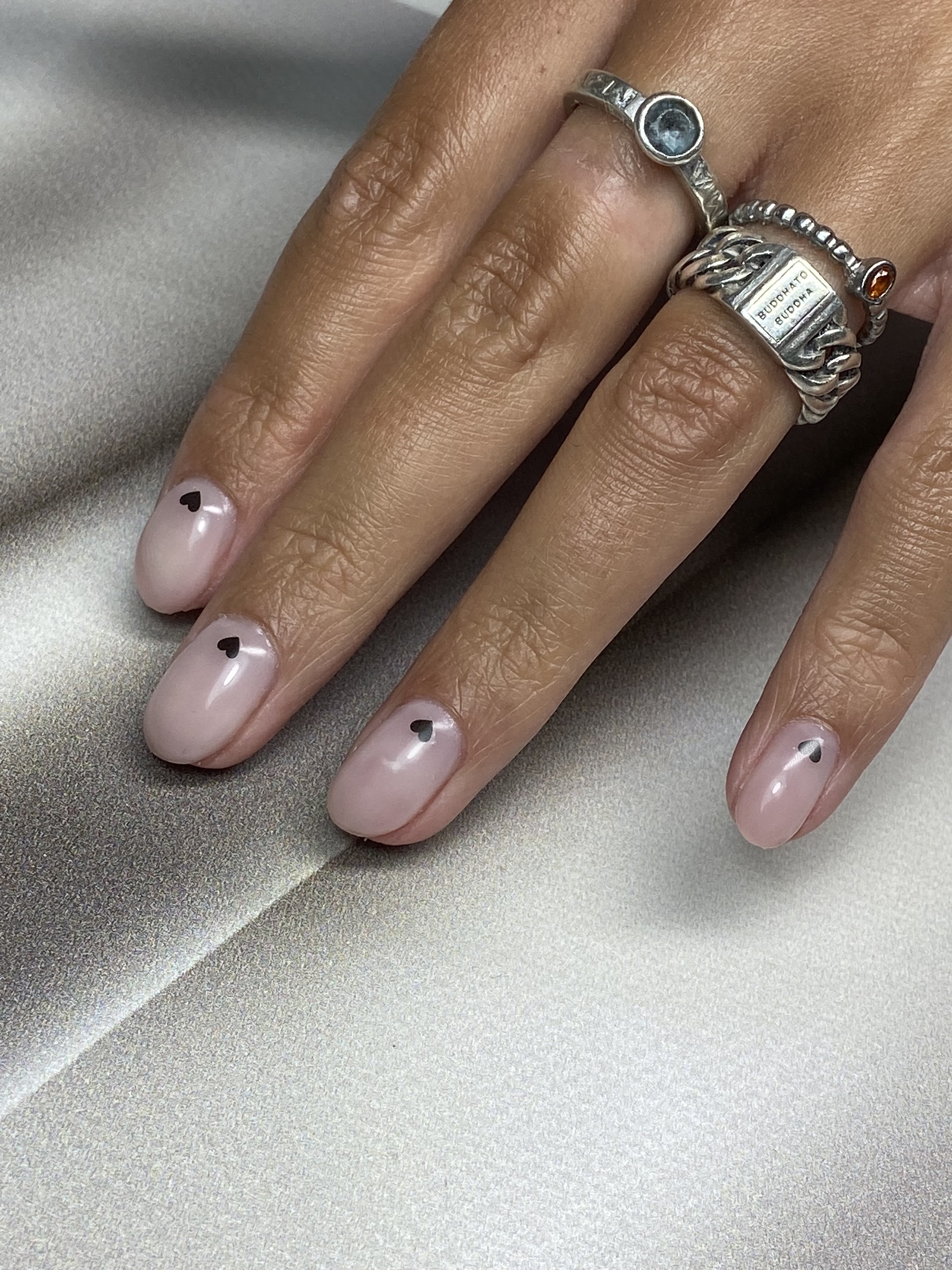 Nude nagels met een klein donker accent per nagel, hand met zilveren ringen op grijs satijn